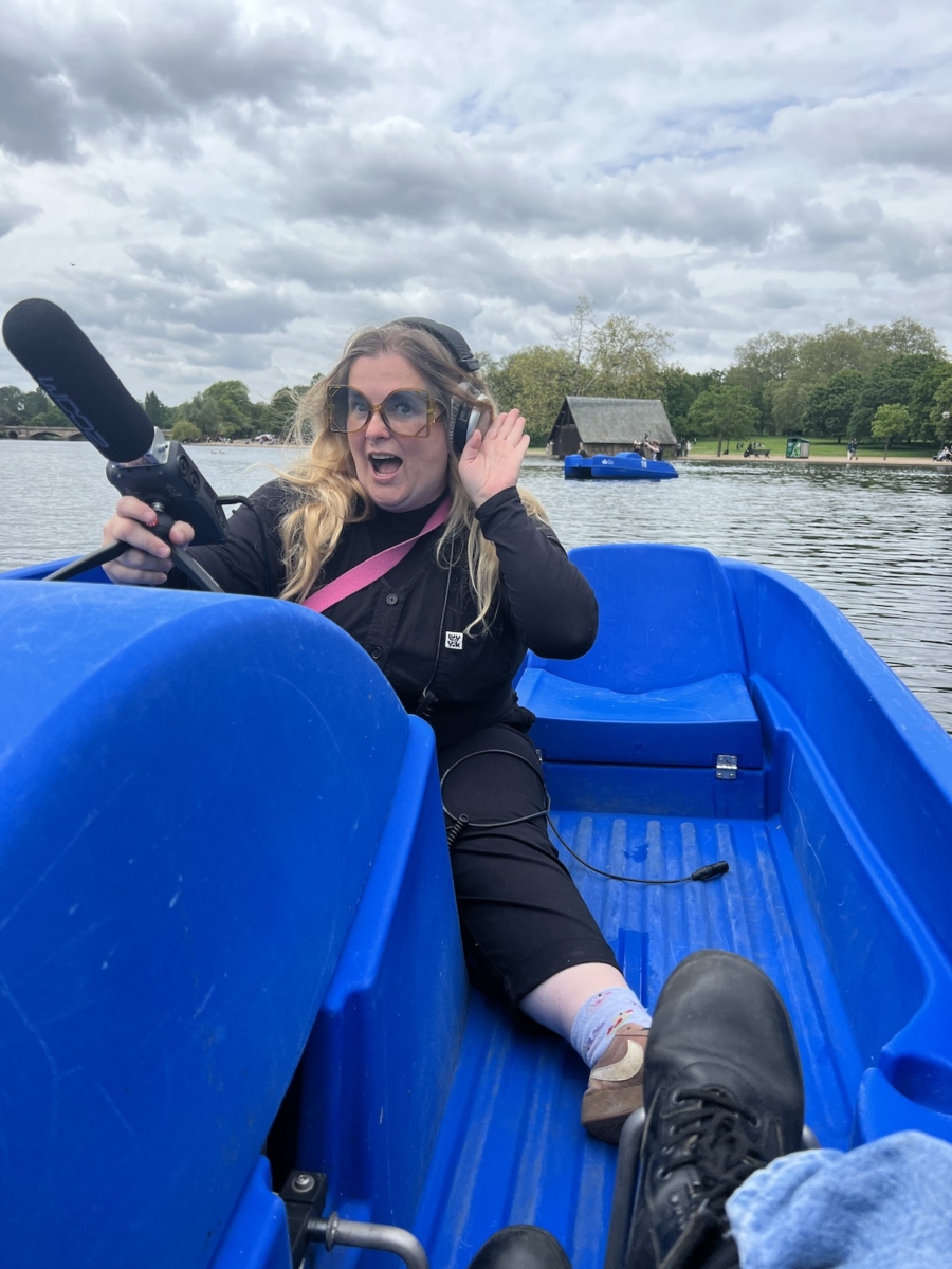 Sounds of the Subaquatic in The Royal Parks, London, led by sound artist Emily Peasgood, 2023. Emily Peasgood is sat on a pedalo in The Serpentine at Hyde Park. She is hold a shotgun microphone, and her other hand is cupping her ear as if to say: "Listen". She has very large glasses that make her look like a bug, and is smiling broadly.