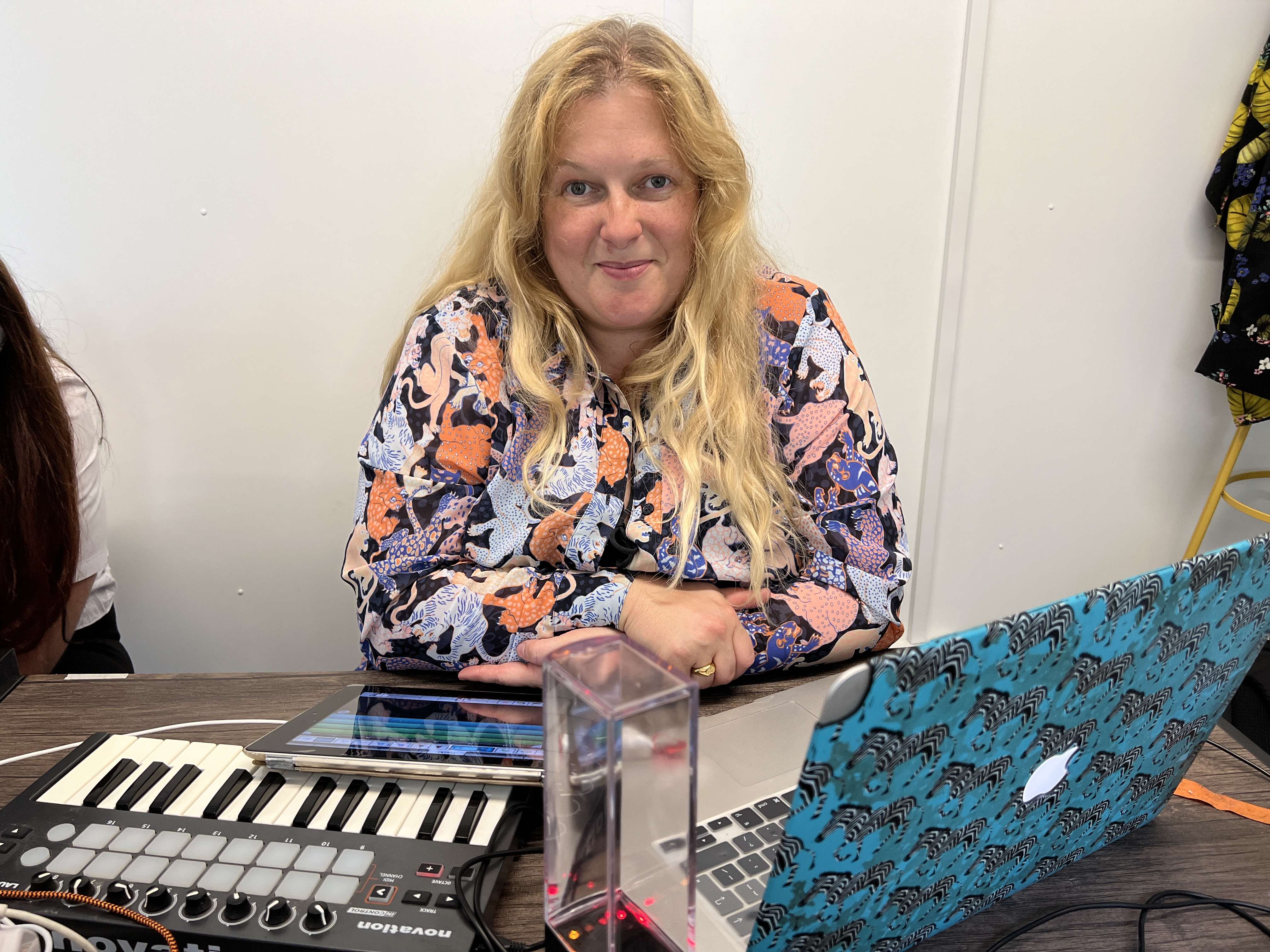 This image shows lead artist Emily Peasgood sat at a table in the comunity hub at Victoria Park in Ashford. She is smiling at the camera and a small MIDI keyboard, IPAD and laptop are positioned in front of her. 