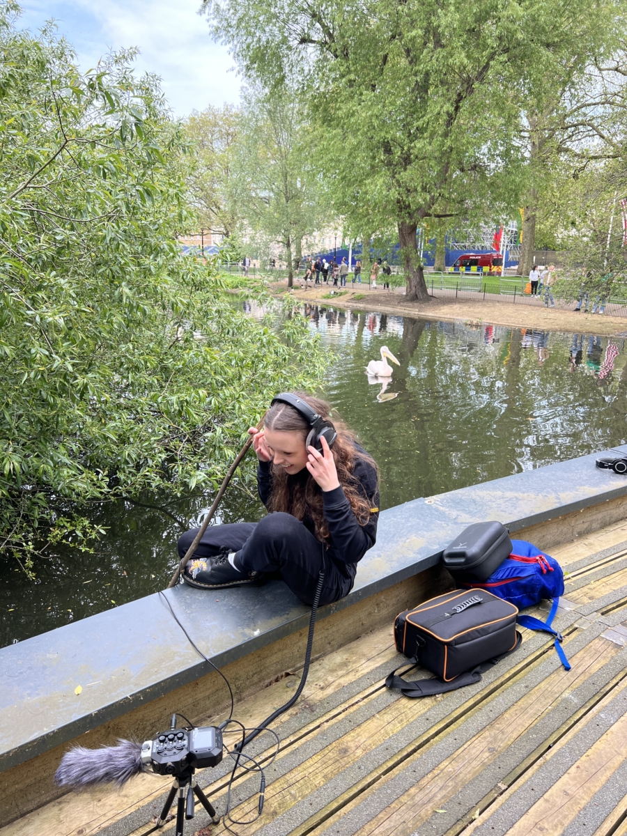Sounds of the Subaquatic in The Royal Parks, London, led by sound artist Emily Peasgood, 2023. Eleanor Fineston-Roberts (student at Guildhall School of Music and Drama) sits on the bank of St James's Lake in St James's Park. She is wearing headphones and smiling as she listens to the sounds she has recorded.