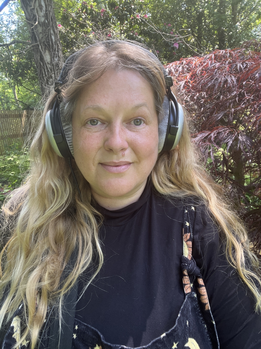 Sounds of the Subaquatic in The Royal Parks, London, led by sound artist Emily Peasgood, 2023. Emily Peasgood is wearing headphones and smiling at the camera.