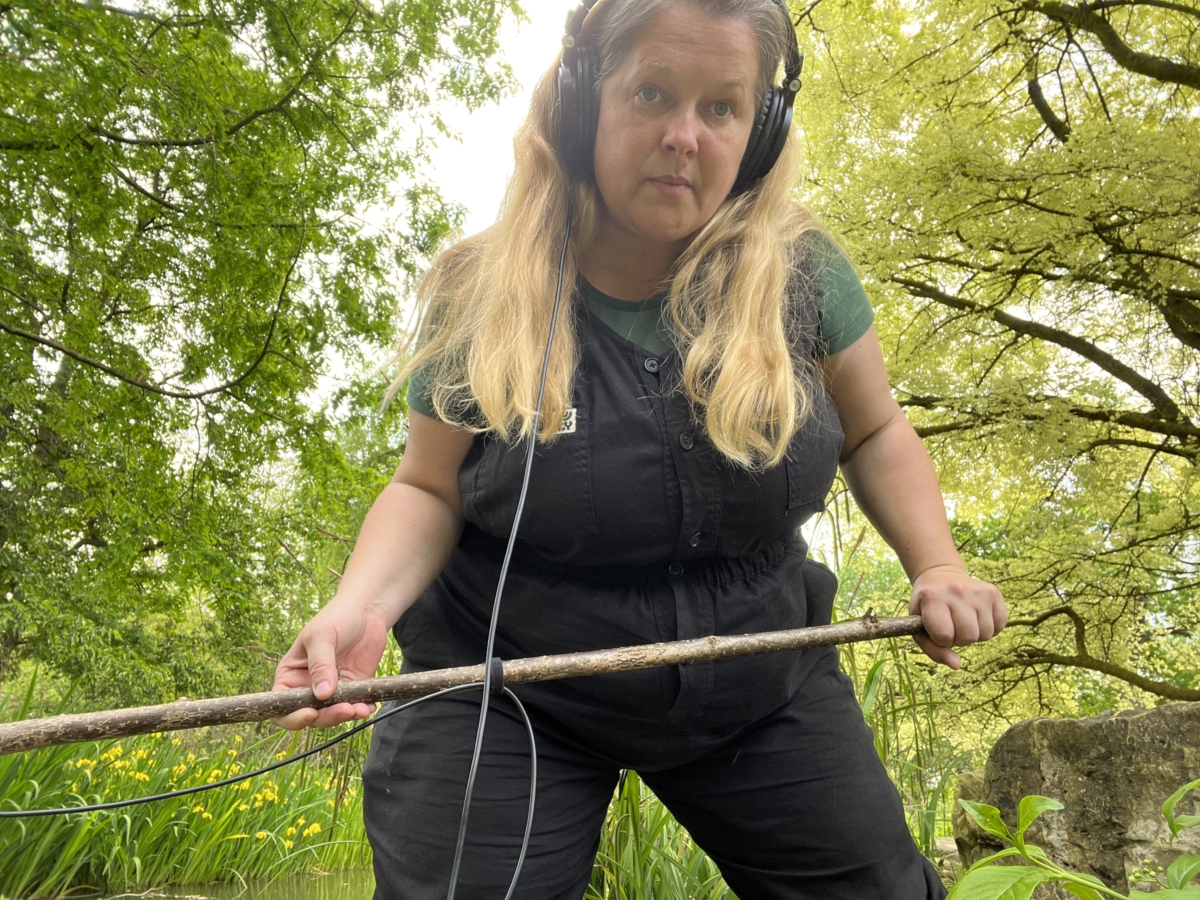 Sounds of the Subaquatic in The Royal Parks, London, led by sound artist Emily Peasgood, 2023. Emily Peasgood is looking at the camera. She is holding a long birch branch with a hydrophone attached, and wearing headphones.