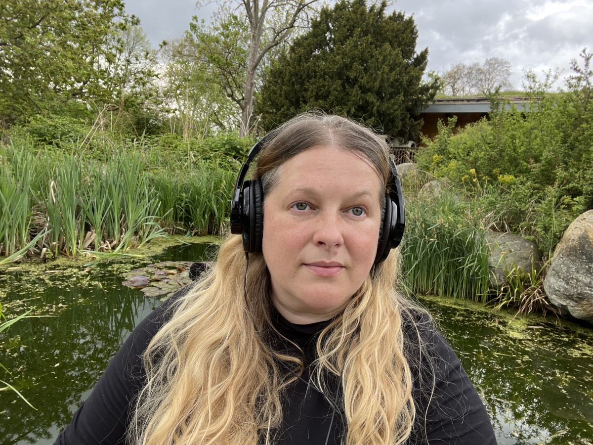 Sounds of the Subaquatic in The Royal Parks, London, led by sound artist Emily Peasgood, 2023. Emily Peasgood stands next to the pond at The LookOut Garden in Hyde Park. She is wearing a large pair of headphones and staring thoughtfully at the camera.