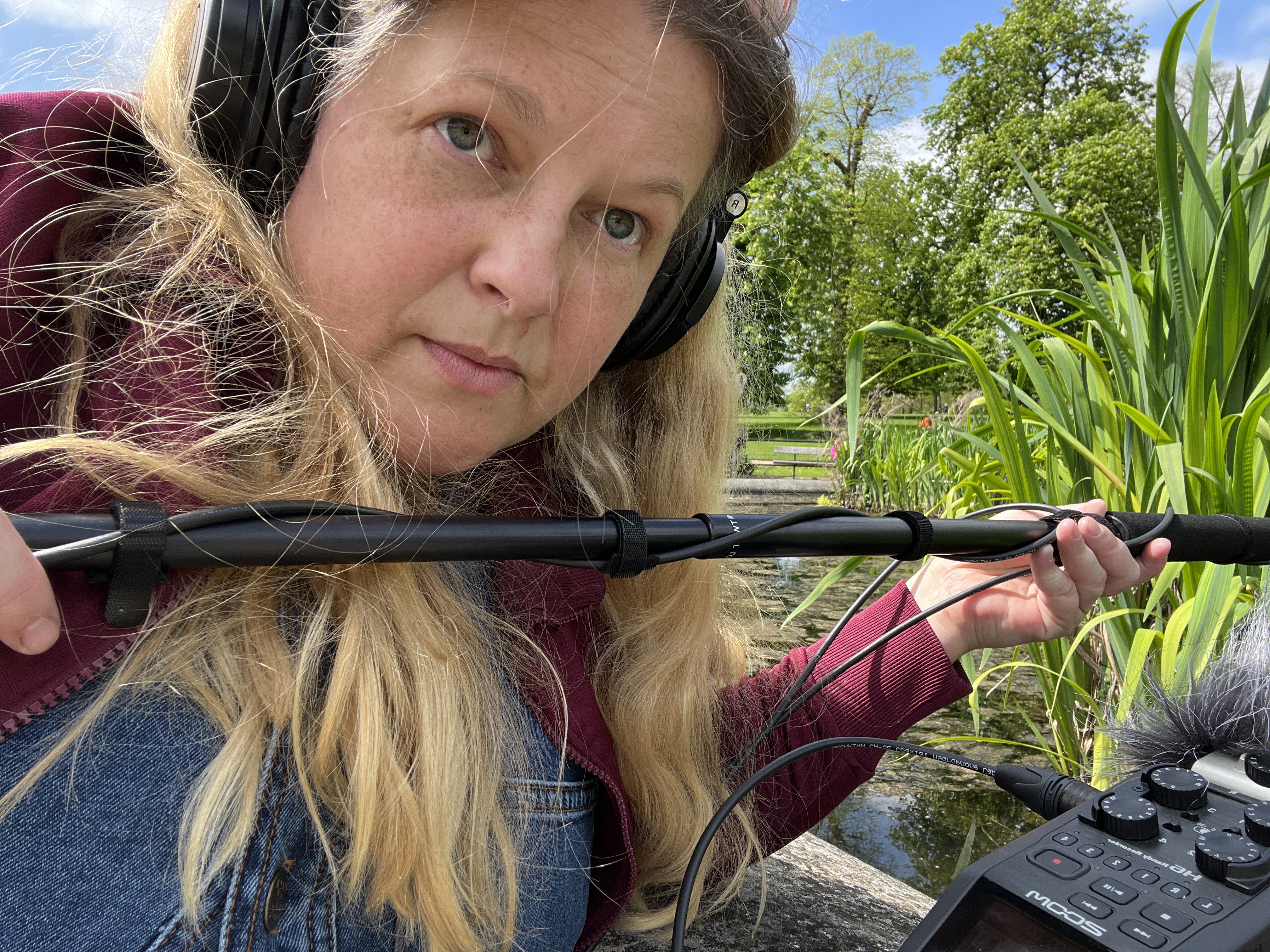 Sounds of the Subaquatic in The Royal Parks, London, led by sound artist Emily Peasgood, 2023. Emily Peasgood is holding a boom pole with a hydrophone attached and wearing headphones. She is sat next to a pond in The Italian Gardens at Kensington Gardens. 