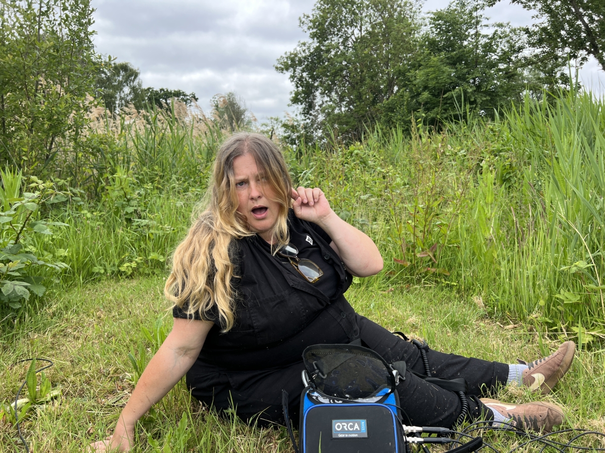 Sounds of the Subaquatic in The Royal Parks, London, led by sound artist Emily Peasgood, 2023. Emily Peasgood is sat on some grass in The Regents Park. She is pulling a silly face and motioning to her ear to encourage the photographer to listen.