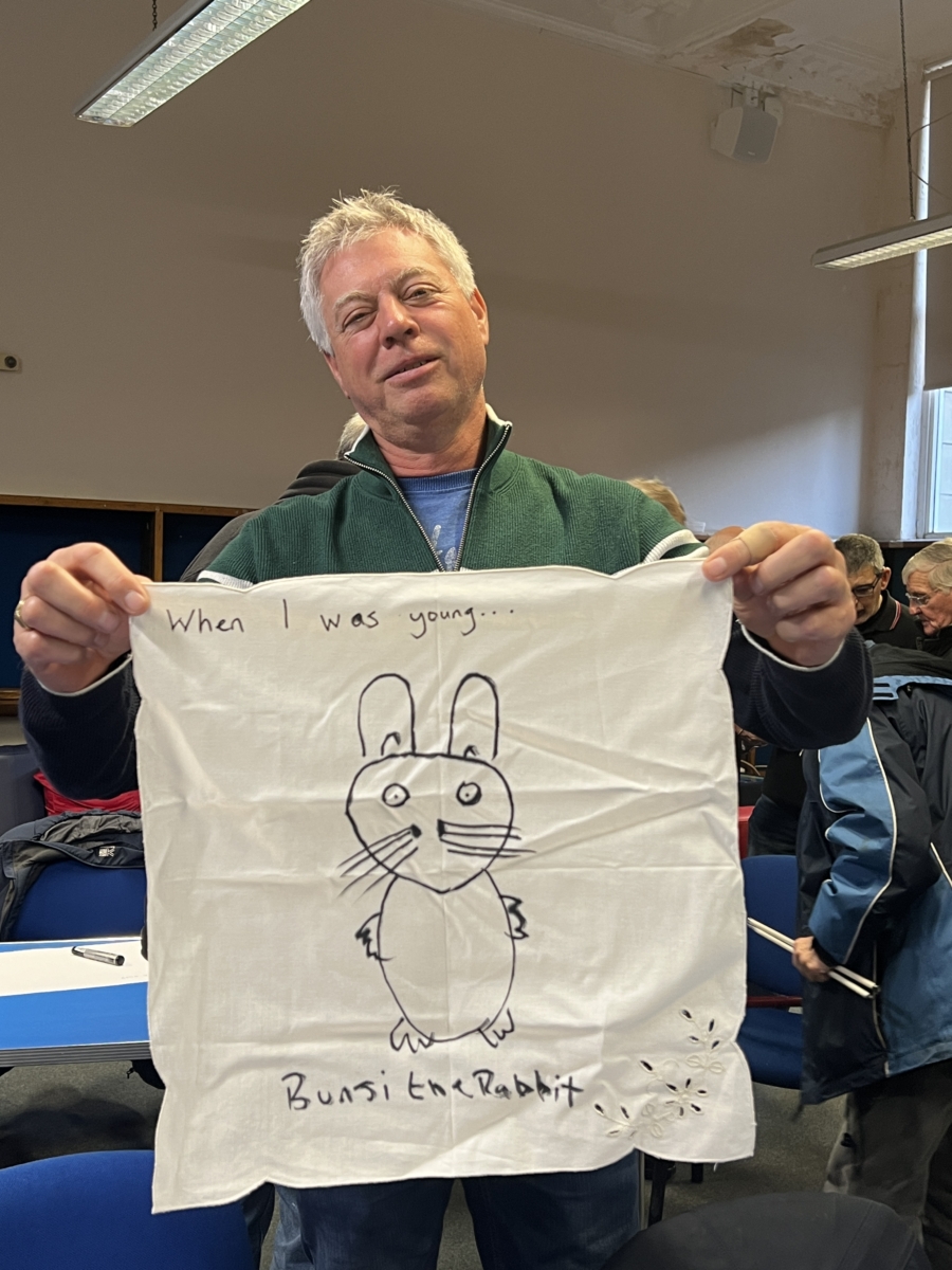A man holds up a piece of laundy he has written on. It shows a picture of a cuddly rabbit toy and says "Bunsi the rabbit" underneath.