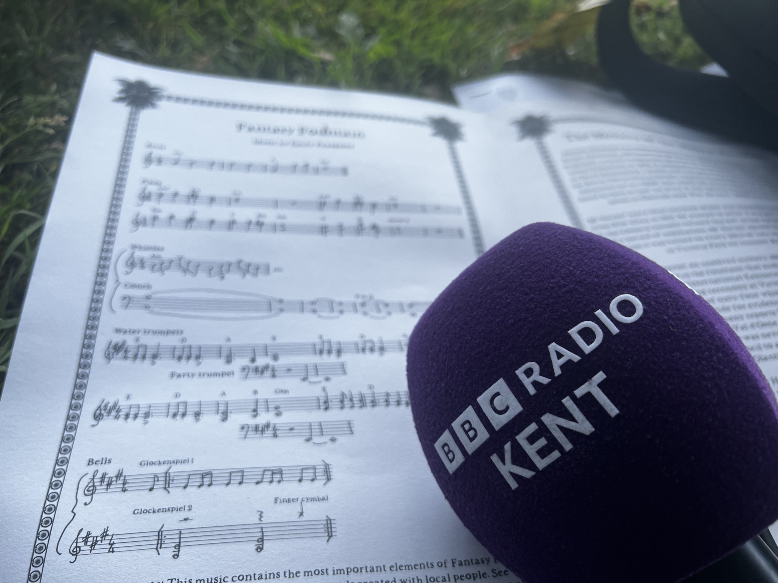 This image shows the limited edition score of Fantasy Fountain. It is placed on the grass in Victoria Park, at the opening day in 2022, and a purple BBC Radio Kent microphone is placed over the top.
