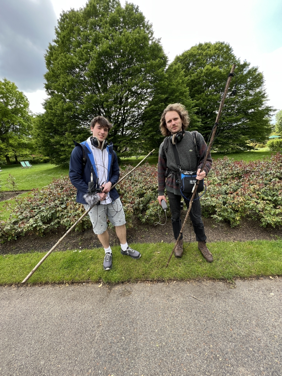 Sounds of the Subaquatic in The Royal Parks, London, led by sound artist Emily Peasgood, 2023. Guildhall School of Music and Drama student James Landsdowne is stood with assistant field recordist Geoffrey Papin, facing the camera with a flower bed in Queen Mary's Garden Pond at The Regent's Park in the background. They are holding long birch poles with hydrophones wrapped around them.