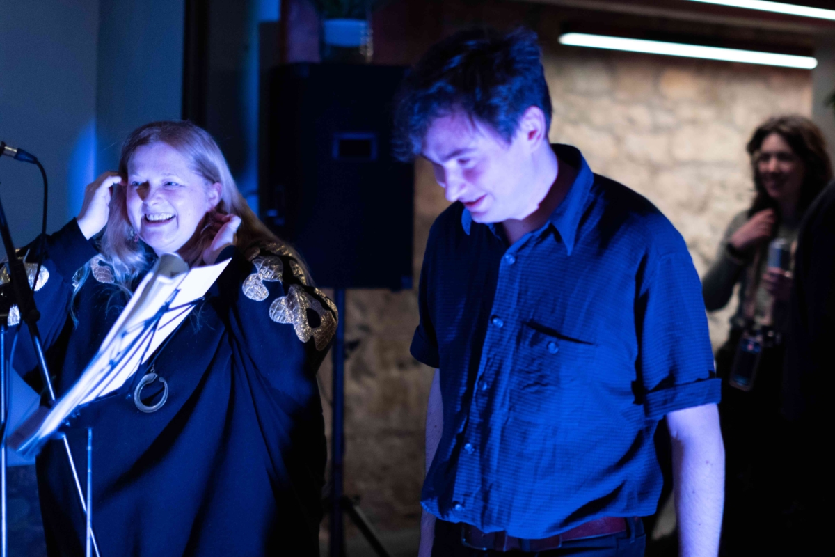 Emily Peasgood and musician Benji Heinke are standing on the stage in the God's House Tower bar, at the launch event of When I Grow Up I Want To Be A Ship. They are both smiling. 
