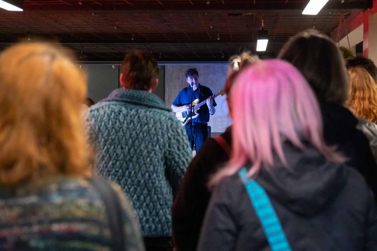 This image is taken from the back of the bar at God's House Tower and shows the backs of several members of the public's heads. In the distance, musician Benji Heinke can be seen singing his interpretation of When I Grow Up I Want To Be A Ship at the launch event in 2022.
