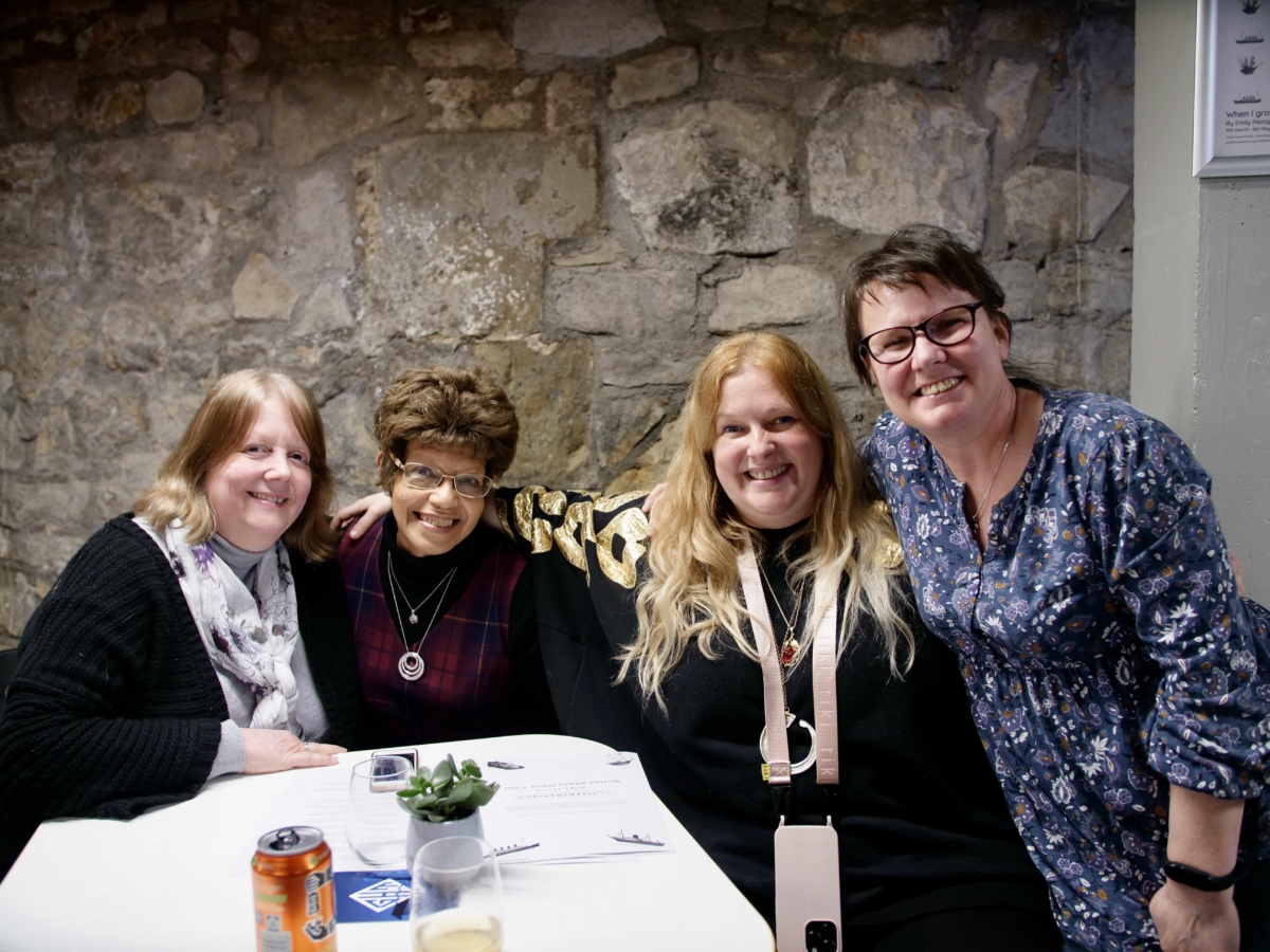 Sandra Peasgood, Pamela Ward, Emily Peasgood and co-lyricist of 'When I Grow Up I Want To Be A Ship' Kate Lynne-Devere are sitting around a table in the bar of God's House Tower, at the launch event of When I Grow Up I Want To Be A Ship in 2022. They have their arms around each other and are smiling at the camera.
