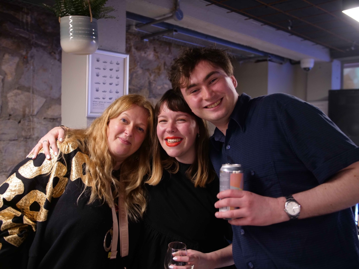 Artist Emily Peasgood stands with Creative Programme Manager Mia Delve and musician Benji Heinke at the opening even of When I Grow Up I Want To Be A Ship in the bar of God's House Tower, 2022. They all smile at the camera. 