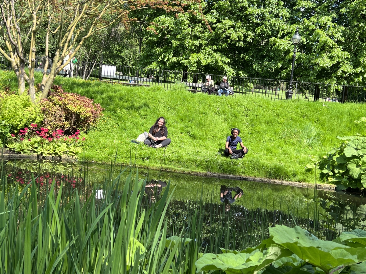 Sounds of the Subaquatic in The Royal Parks, London, led by sound artist Emily Peasgood, 2023. Rebecca Huxley (guest artist) and Dani Osoba (assistant field recordist) are sat on a river bank in Hyde Park, while they record sounds of the Dell Stream with hydrophones.