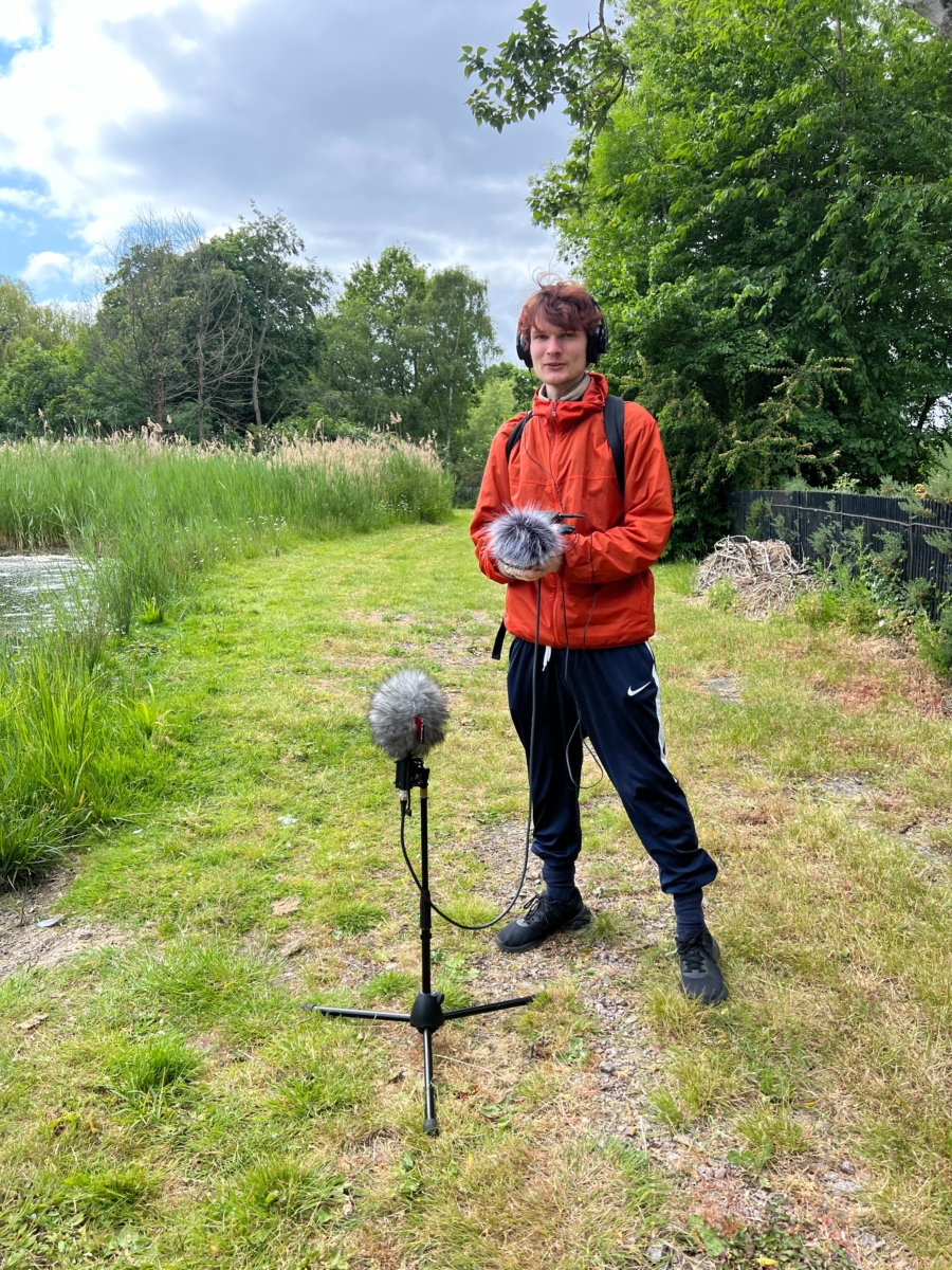 Sounds of the Subaquatic in The Royal Parks, London, led by sound artist Emily Peasgood, 2023. Seamus Heath (Guildhall School of Music and Drama student) is standing next to a wetland pond in The Regent's Park. He is holding recording equipment and smiling at the camera. 