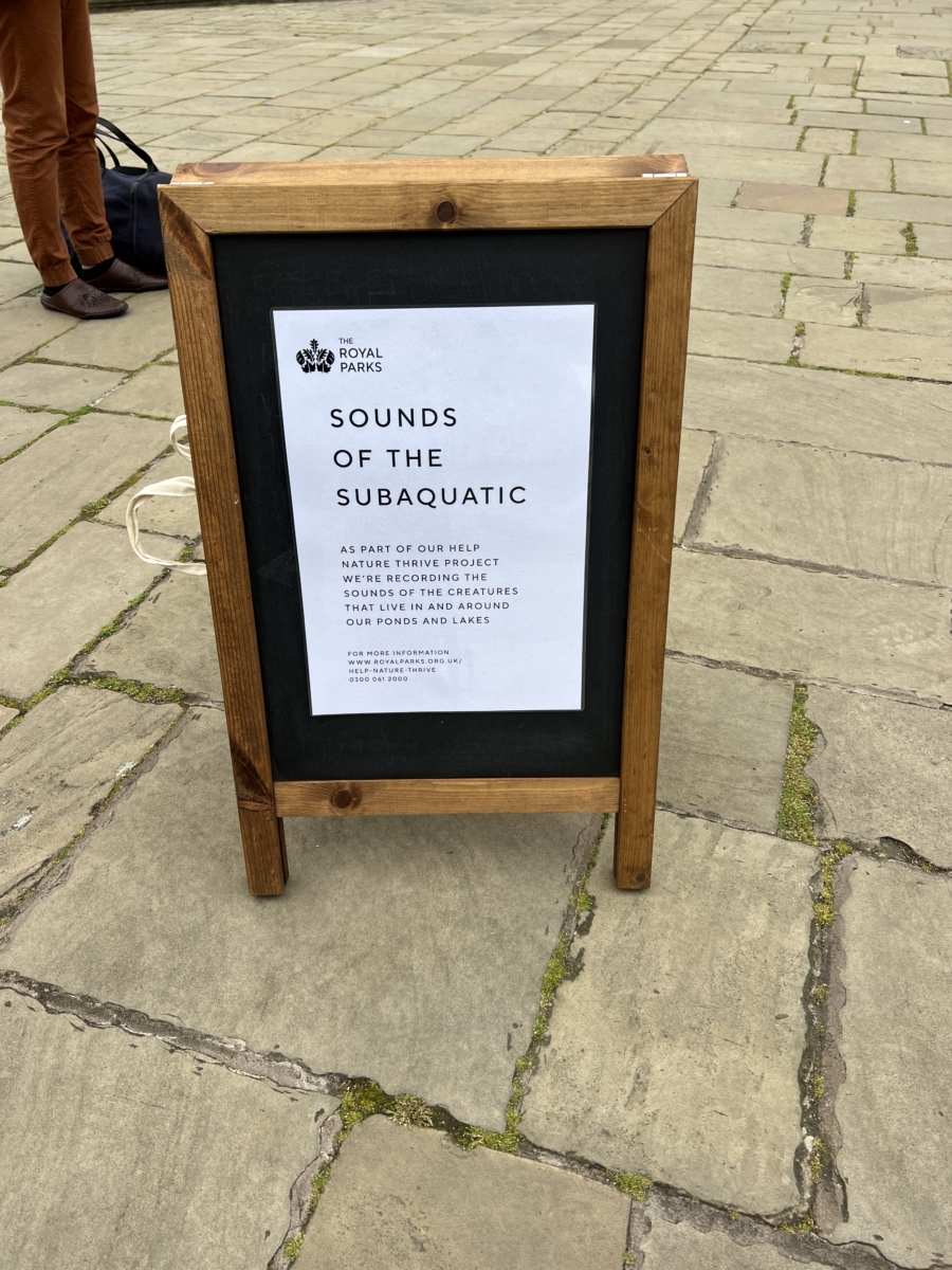 Sounds of the Subaquatic in The Royal Parks, London, led by sound artist Emily Peasgood, 2023. A wooden A-frame stands on the ground in The Italian Gardens at Kensington Gardens. It has a poster attached stating: "Sounds of the Subaquatic". 