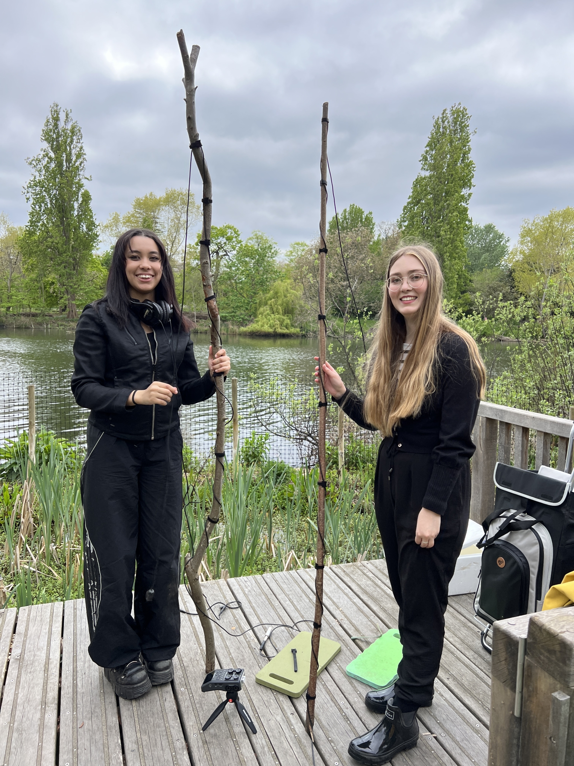 Sounds of the Subaquatic in The Royal Parks, London, led by sound artist Emily Peasgood, 2023. Guildhall School of Music and Drama students Tilley Gartan and Isabel Woodings are standing on a wooden platform on The Long Water in Kensington Gardens. They each hold a long birch branch with a hydrophone wrapped around it and are smiling at the camera.